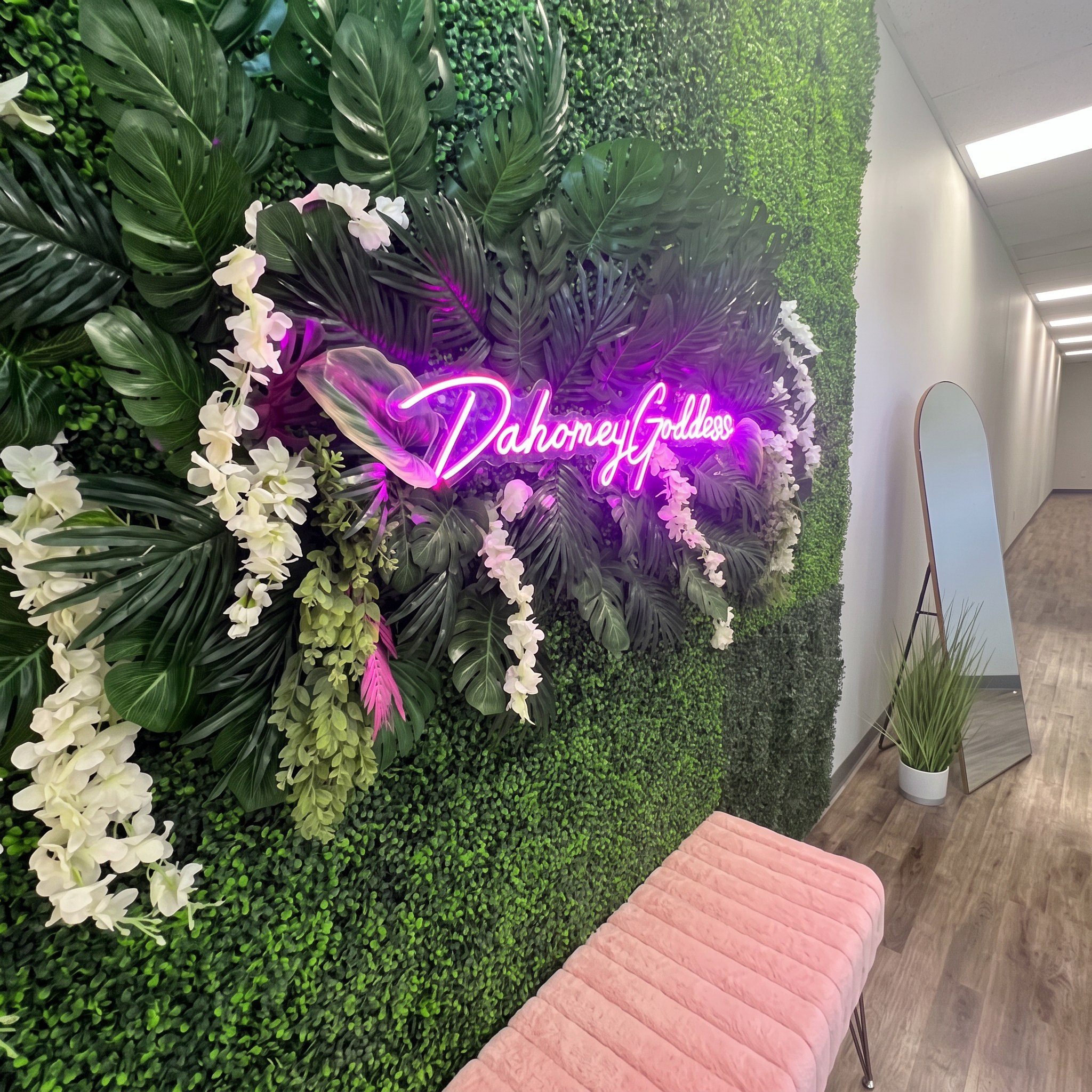 Dahomey Goddess neon sign on a lush greenery wall with a pink velvet bench at the Greenville, NC studio